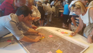 Crowded room with people actively involved with the engagement process writing notes on post-it stickers and placing on large maps