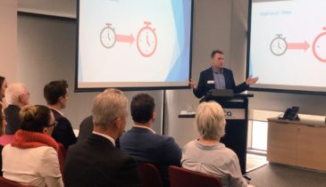 Photo of a presentation with people seated listening to speaker